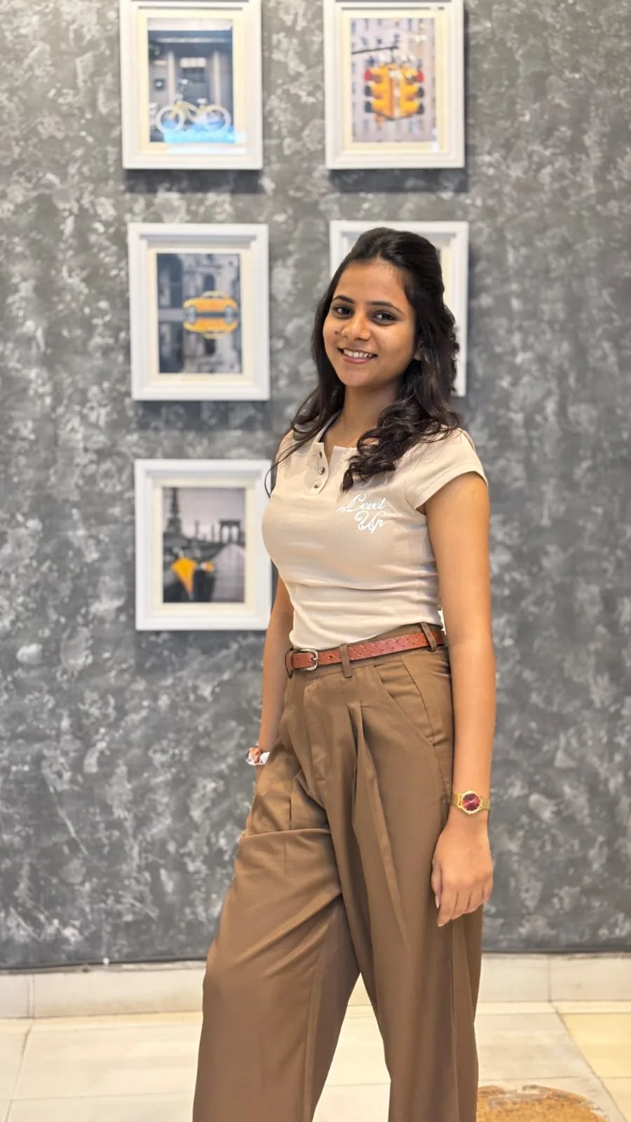 Megha Choudhary standing in a beige top and brown trousers beside framed photographs